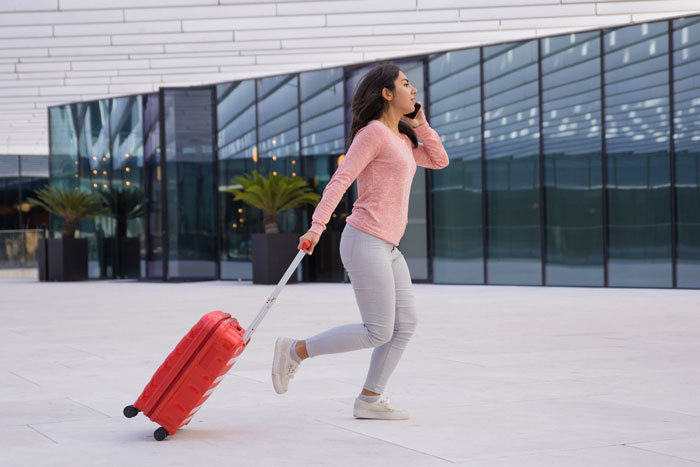 Young woman with a red suitcase running outside a modern building, looking concerned and distracted on her phone.