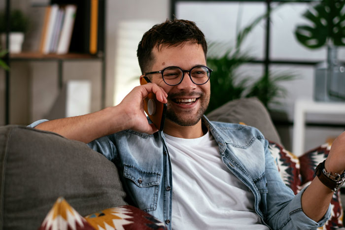 Man laughing on phone call while sitting on couch in a cozy living room after a boyfriend's dumb prank gone wrong.