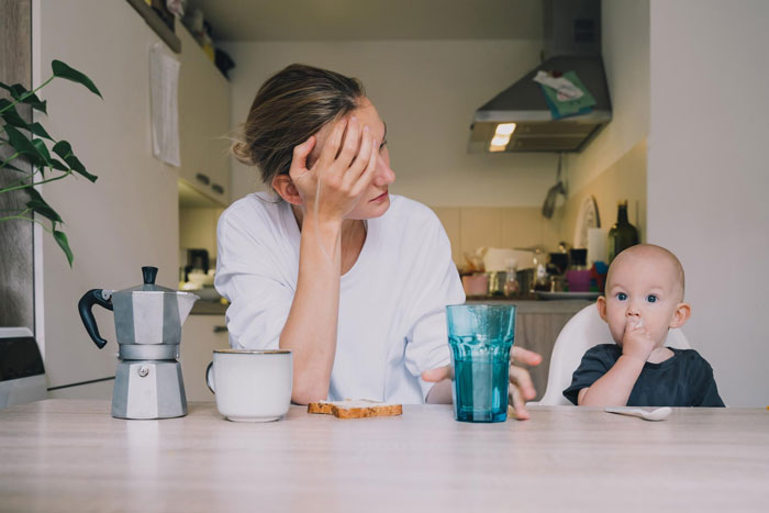 Pregnant sister sitting at kitchen table with baby, looking stressed and tired in a modest homeless shelter setting.