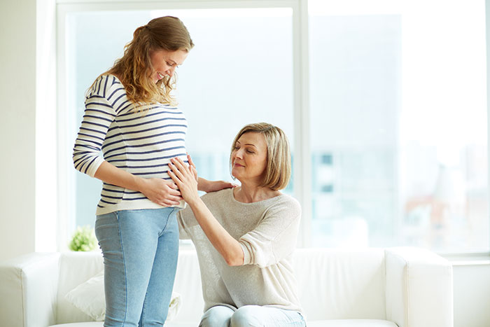 Pregnant woman uncomfortable with mother-in-law actions touching her belly while sitting on a white couch in bright room