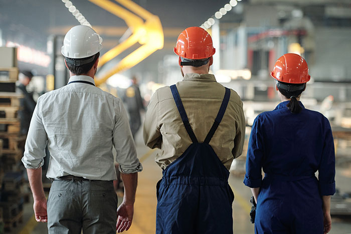 Three factory workers wearing safety helmets stand with backs to camera in industrial workplace, illustrating coworkers getting fired fast.