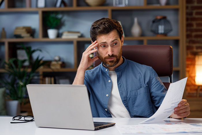 Man in casual office setting looking stressed while reviewing documents near laptop, illustrating coworkers getting fired fast.