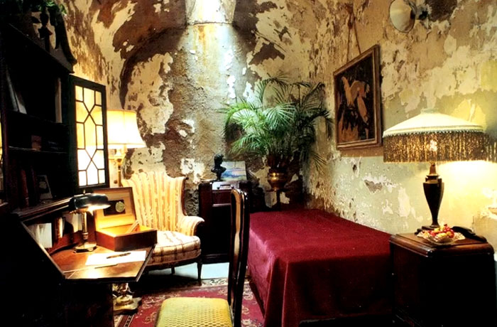 Historic room with worn walls, vintage furniture, and a red-covered bed showcasing rare and intriguing photos of history.