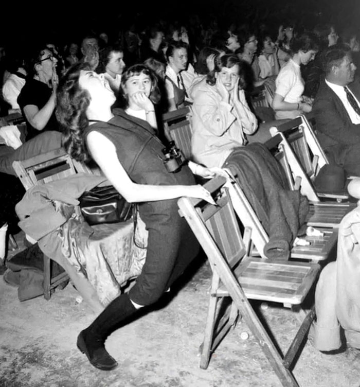 Black and white rare history photo showing a woman leaning back on a chair in a crowded indoor event setting.