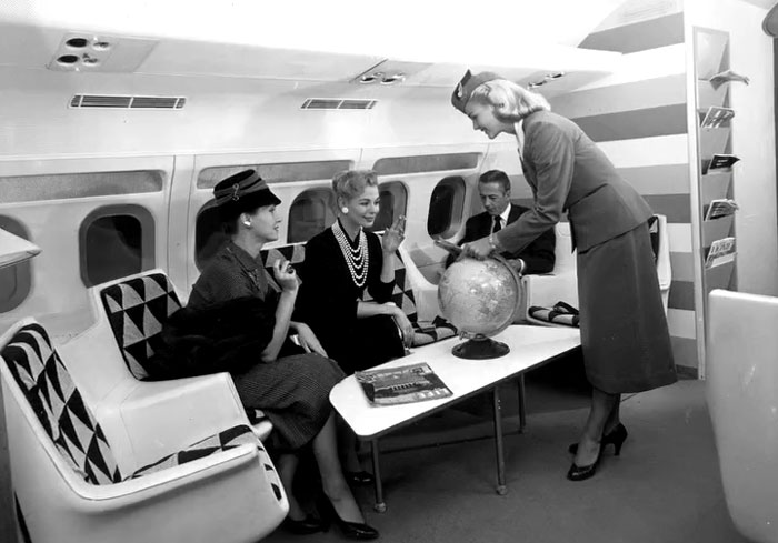 Vintage photo showing airline passengers and a flight attendant interacting with a globe inside an airplane cabin.
