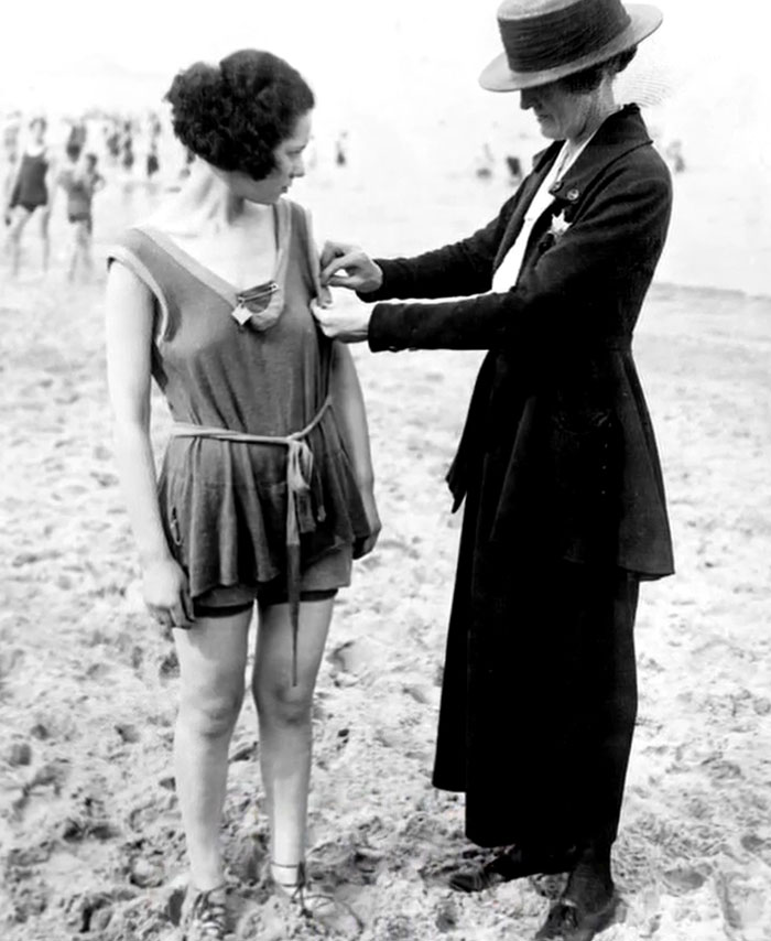Black and white rare historical photo of a woman pinning a badge on a young woman’s bathing suit on the beach.
