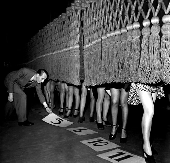Man inspecting numbered cards on floor while women’s legs are visible under fringed curtain in rare history photo.