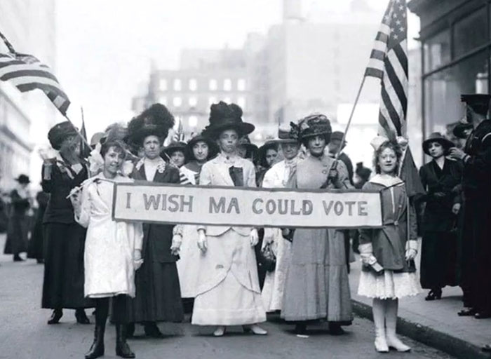 Group of women in vintage clothing holding a banner and American flags in a rare historical photo showing history between headlines.