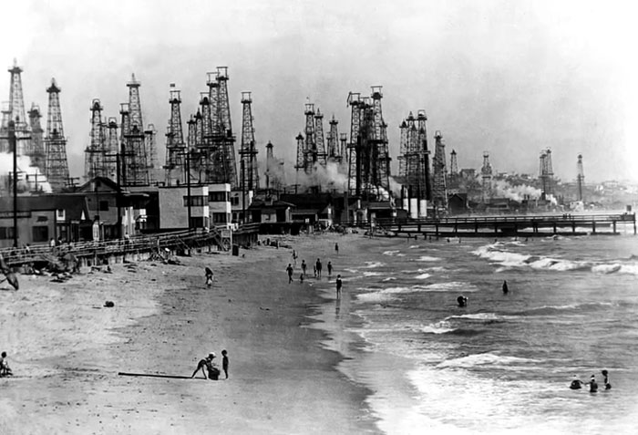 Black and white photo of people on a beach with numerous oil rigs in the background, rare historical scene.