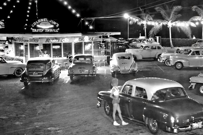 Black and white photo showing rare and intriguing historic cars and people outside Colonel Jim’s restaurant at night.