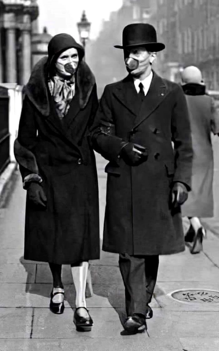 Couple wearing early 20th century gas masks walking on city street, rare and intriguing historical photo.