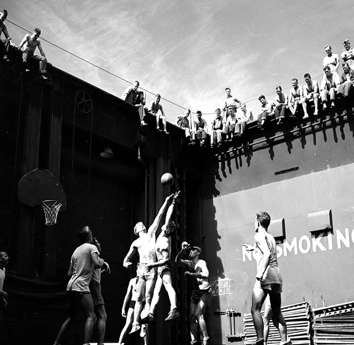 Black and white rare photo of young men playing basketball with others watching from above, showing history between the headlines.