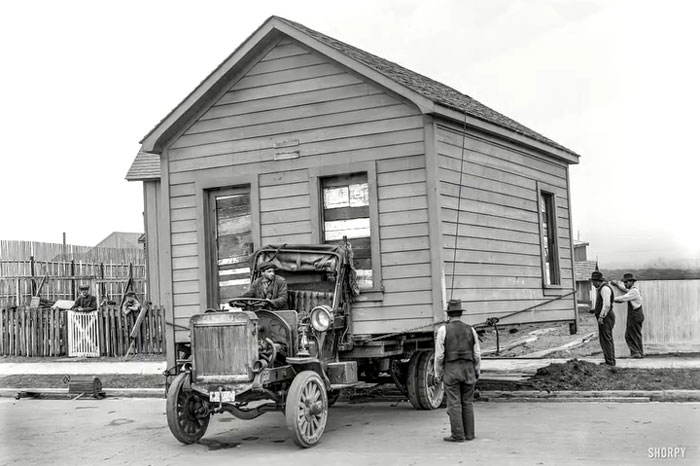 Vintage photo showing rare historical moment of a large house being moved on an early 20th-century truck on city street.