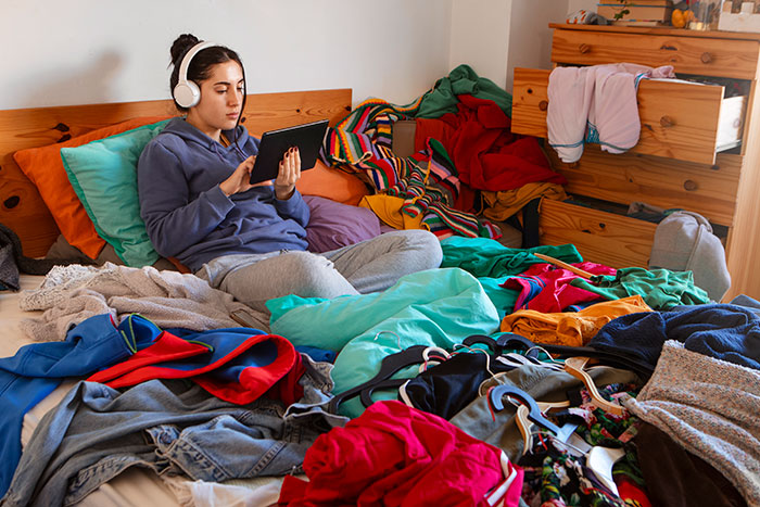 Young woman wearing headphones using tablet in a cluttered bedroom filled with messy clothes, showing red flags in a home.