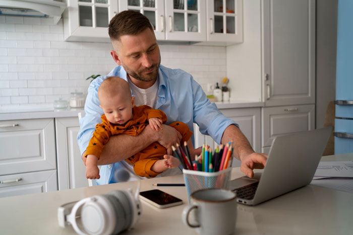 Man holding baby while working on laptop in kitchen, representing 50YO dad and surprise baby news story.