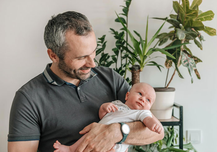 Middle-aged dad holding newborn baby, smiling gently in a bright room with green plants in the background.