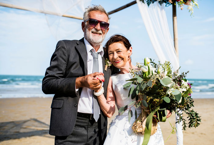 Older couple smiling at beach wedding, bride holding bouquet, highlighting daughter refusing to support dad's wedding cheerleading.