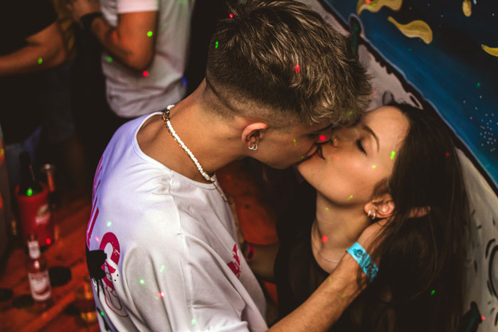 Young couple kissing closely at a party with colorful light spots and casual clothing in a lively setting.