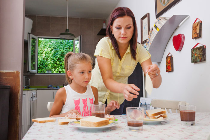 Woman preparing food at table for childfree bestie while child watches, highlighting babysit and feed kids request.