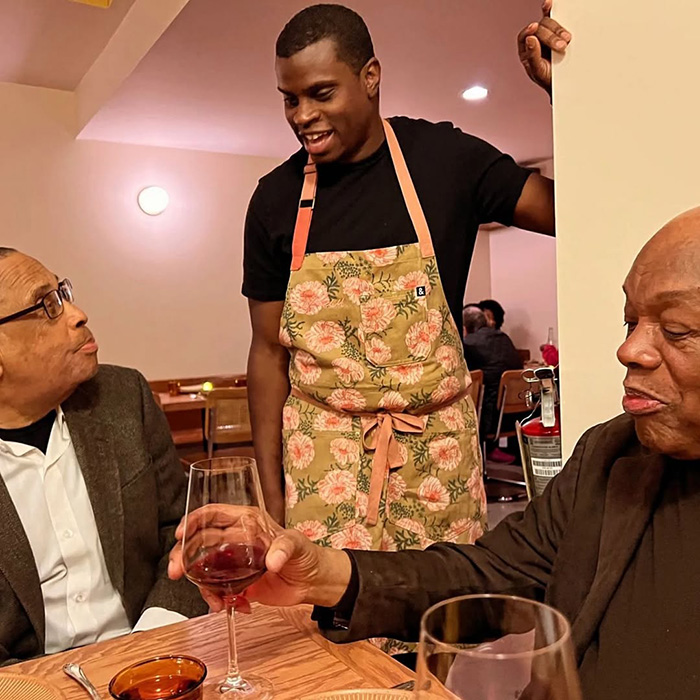 Restaurant server wearing floral apron talks to two seated diners raising a glass amid upset over automatic 20% tip policy.
