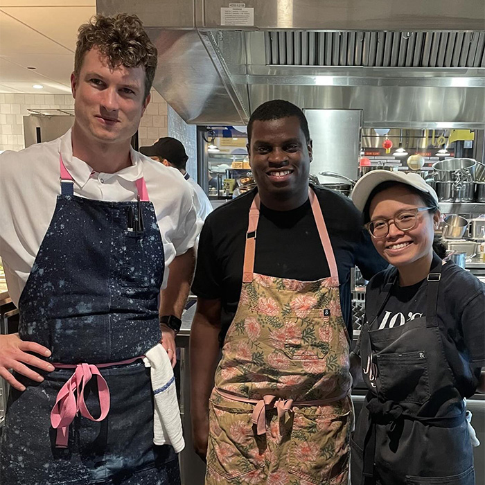 Three restaurant staff wearing aprons pose in a kitchen amid outrage over automatic 20% tip and country's racist past.