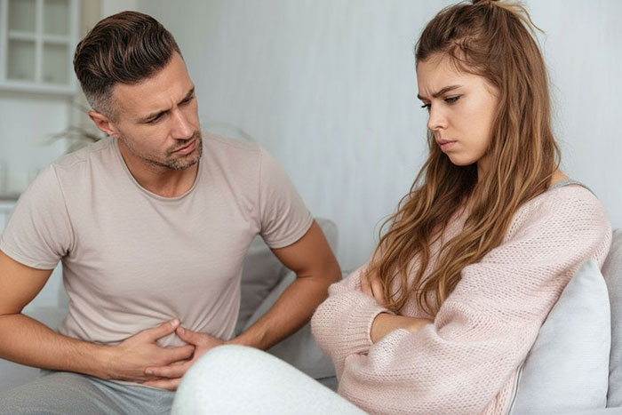 Man holds stomach in pain while fiancée looks upset, highlighting sickness and health challenges in relationships. Man holds stomach in pain while fiancée looks upset, highlighting sickness and health challenges in relationships.