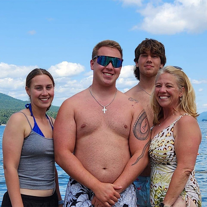 Family of trans mom Roberta Dorgan poses together smiling outdoors near water with blue sky and mountains.