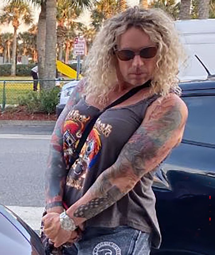 Woman with curly blonde hair and tattoos on her arms wearing sunglasses and a graphic tank top near parked cars outdoors Woman with curly blonde hair and tattoos on her arms wearing sunglasses and a graphic tank top near parked cars outdoors