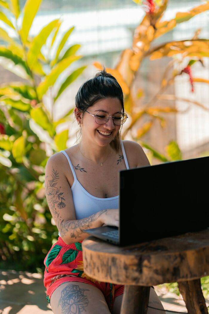 Young woman with tattoos smiling and working on laptop outdoors, related to women list unhinged toxic ways they traumatized men