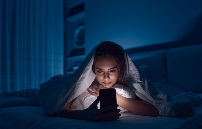 Woman lying in bed under blanket, looking at phone screen with focused expression in dimly lit room at night, RuneScape theme. - 18