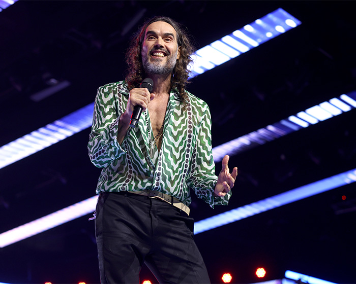 Russell Brand on stage speaking into a microphone, sparking outrage with disturbing charges after hot tub baptisms.