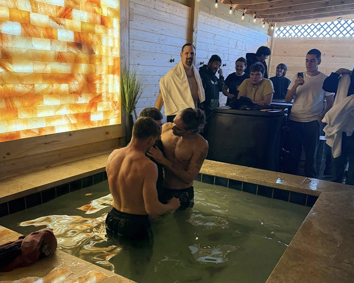 Russell Brand baptizing people in a hot tub while onlookers watch inside a wooden room with warm lighting.