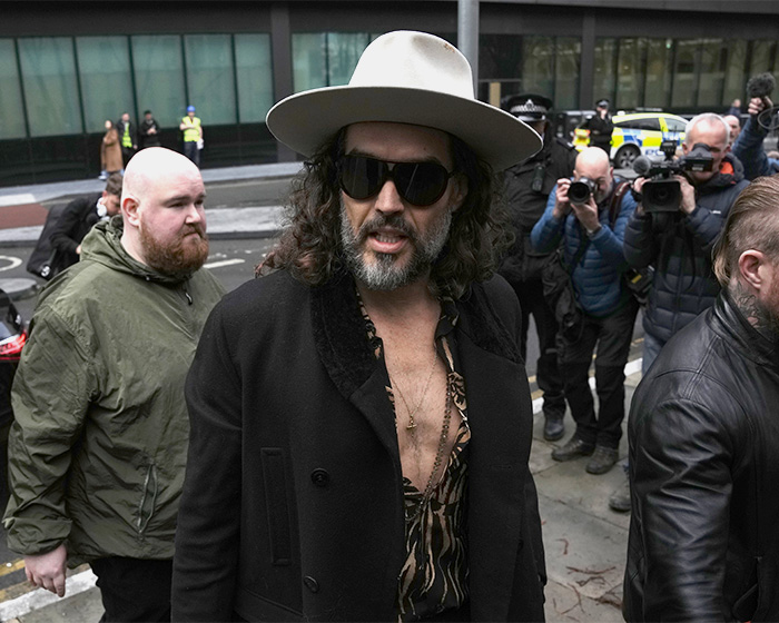 Russell Brand wearing hat and sunglasses, walking past photographers amid outrage over disturbing charges.
