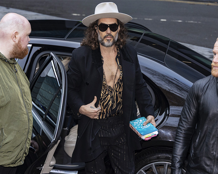 Russell Brand wearing hat and sunglasses, holding a book, stepping out of a car amid public attention and controversy.