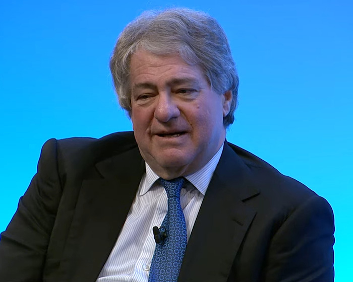 Man in a suit and blue tie speaking during an interview against a solid blue background about Epstein files link. Man in a suit and blue tie speaking during an interview against a solid blue background about Epstein files link.