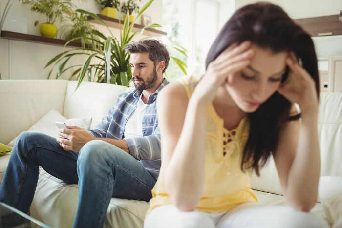 Man focused on phone scheduling hobbies before breakfast, woman in foreground counting minutes feeling ignored at home.