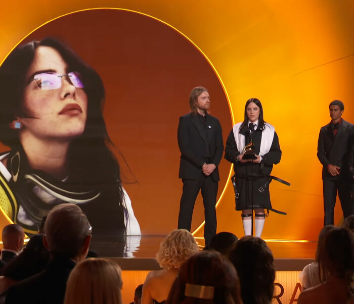 Billie Eilish holding a Grammy on stage with a large portrait behind her during a speech at an awards ceremony.