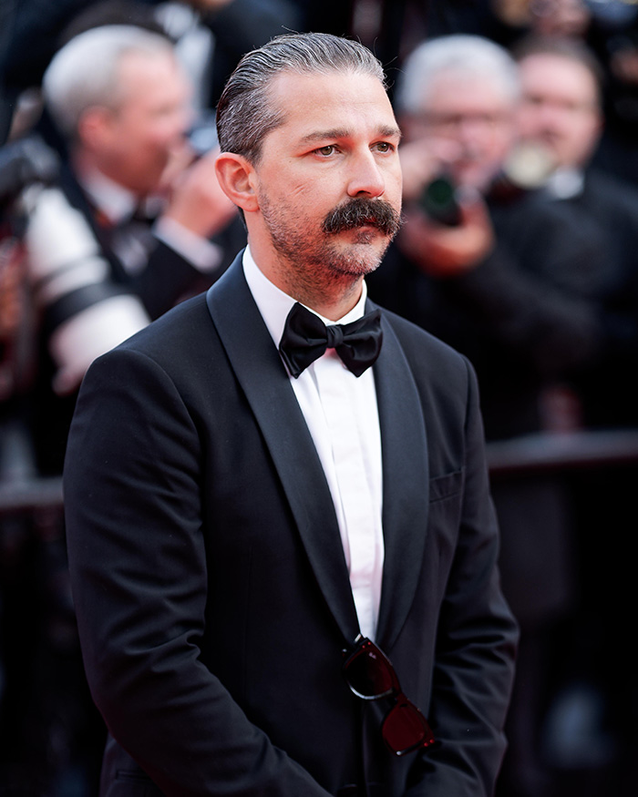 Shia LaBeouf in a black tuxedo with a mustache, attending an event amid a busy crowd with photographers.