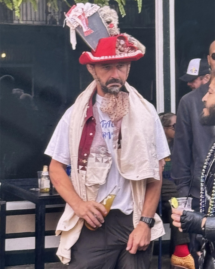 Man in a red hat and layered clothing holding a drink during a street event, illustrating Shia LaBeouf arrest chaos in New Orleans.