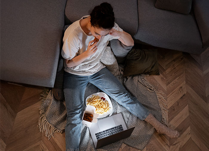 Woman sitting on floor eating fast food while using laptop near couch in a cozy home setting showing signs of low intelligence