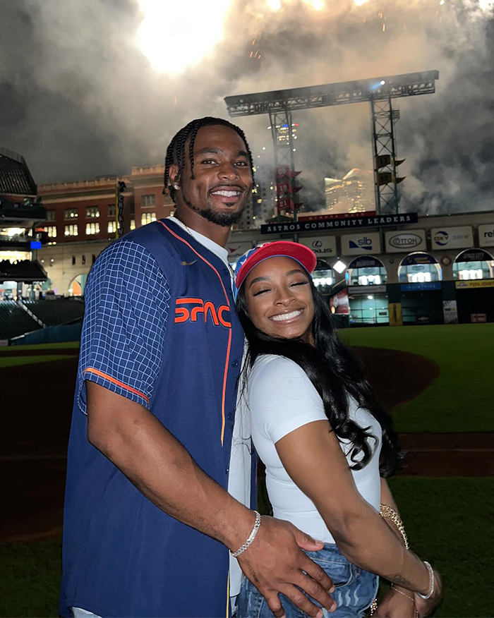 Simone Biles and NFL husband smiling at a stadium with fireworks in the background during the Winter Olympics event.
