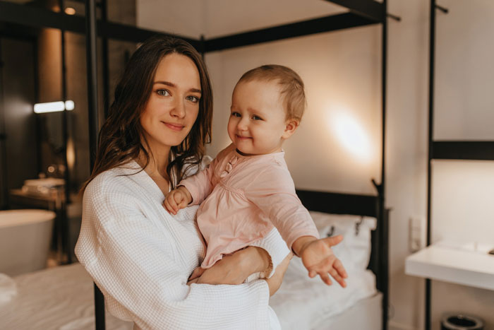 Older sister holding a baby helping single mom in a cozy bedroom, showing care and support with a gentle smile.
