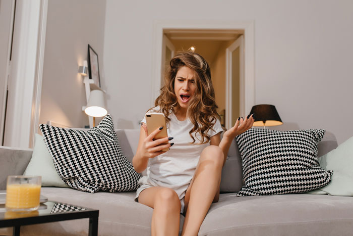 Young woman at home, sitting on a couch, looking frustrated while holding a smartphone, illustrating older sis helping single mom.
