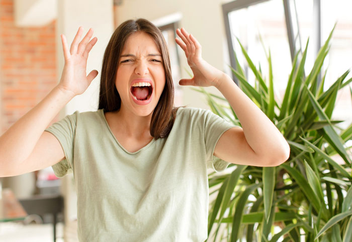 Young woman in casual shirt expressing frustration indoors with green plant and bright window in background, highlighting neglectful mom conflict.