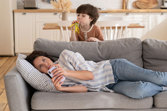 Neglectful mom lying on couch focused on phone while child blows party horn behind her in home setting.