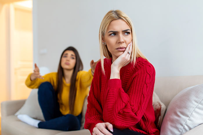 Woman in red sweater looks upset while another woman in yellow gestures from behind on a beige couch, family favor conflict.