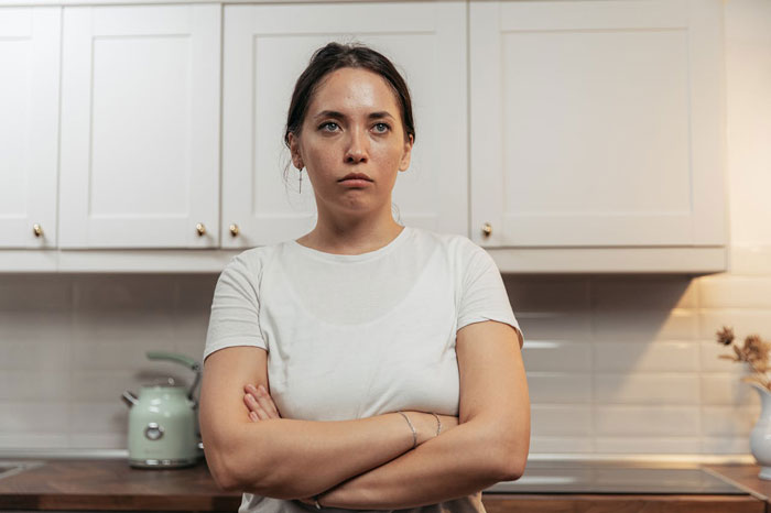 Teen daughter stands with arms crossed in kitchen, showing jealousy and tension related to widowed dad's new love.