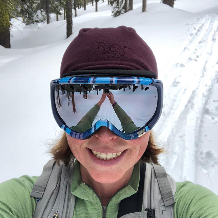 Woman wearing ski goggles and winter hat smiling in snowy forest, related to mothers tragic incident outdoors.