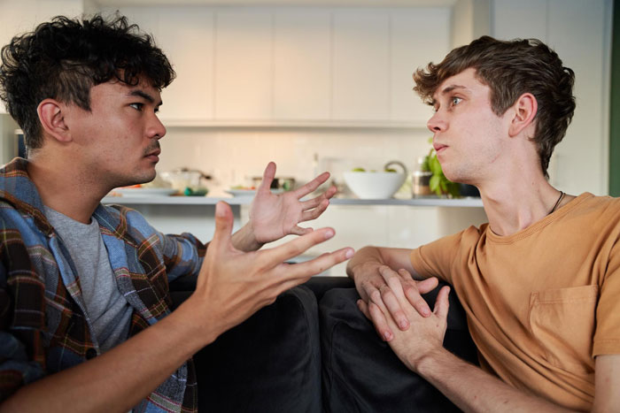 Two young men in a serious discussion on a couch, expressing tension related to girlfriend and best friend conflict.