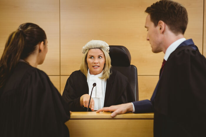 Female judge in traditional wig and robe speaking to two lawyers in a courtroom setting during a legal discussion.